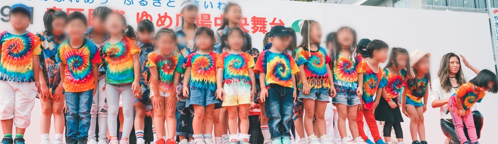 屋外のステージで、タイダイ染めのTシャツを着た子どもたちが一列に並んで踊っている
