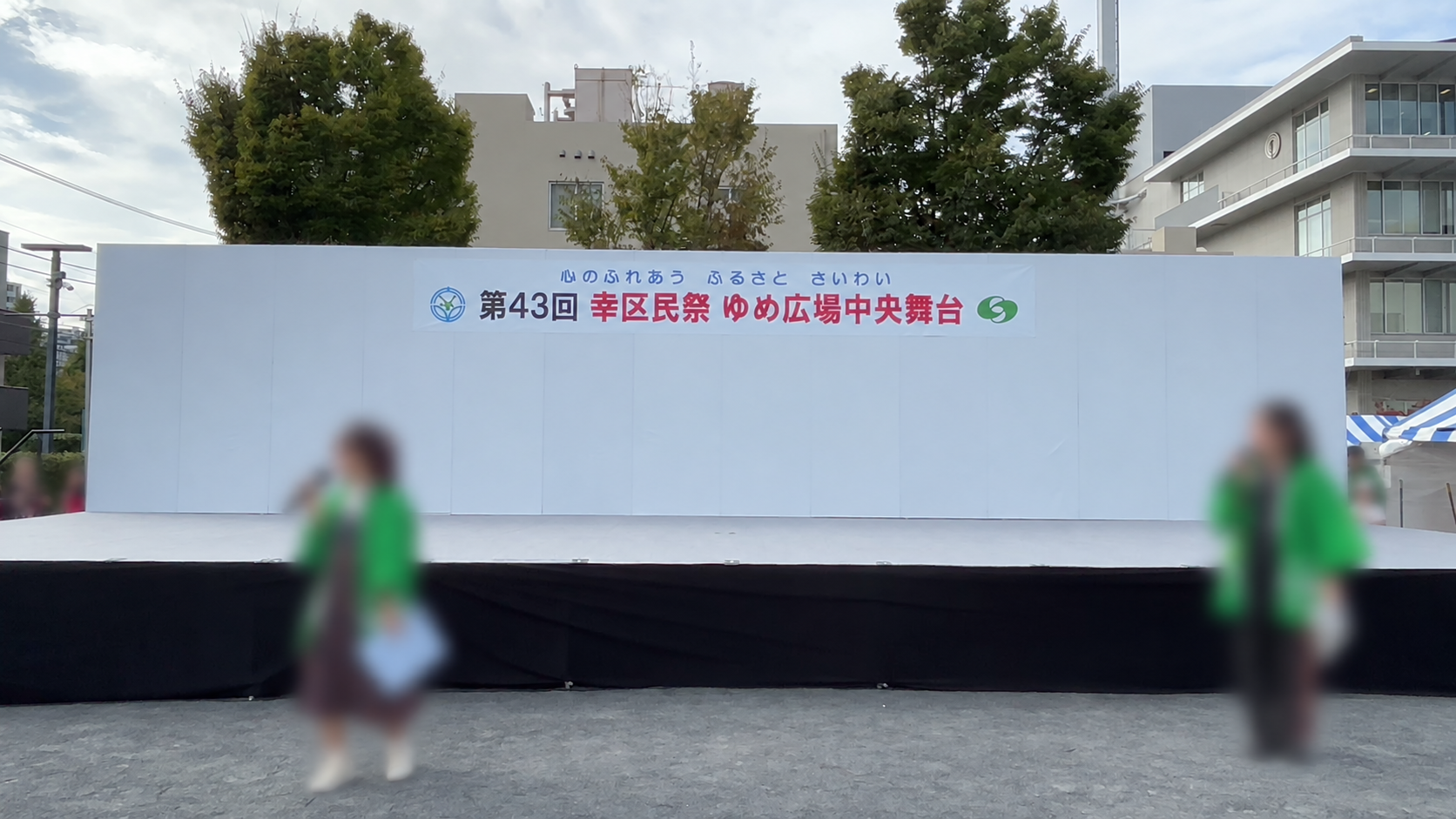 2025年・第43回幸区民祭のゆめ広場中央舞台。白い背面パネルと黒い舞台前面、周囲の木々と建物。人物は識別しにくいよう加工しています。